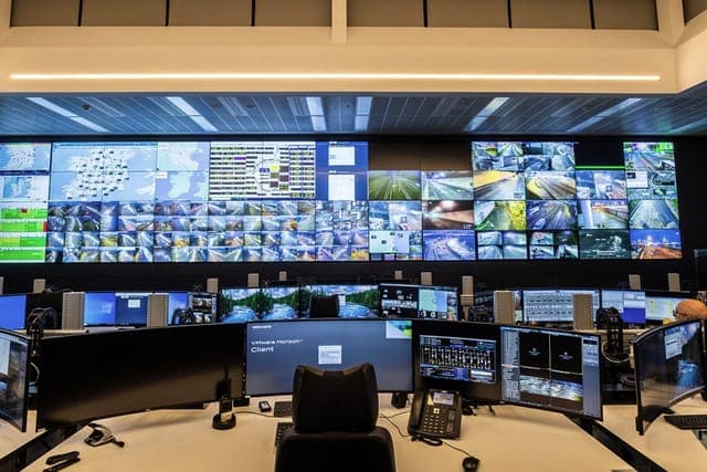 Centro de operações com grande painel de vídeo exibindo mapas e câmeras da rede, sobre uma mesa de comando repleta de monitores onde a equipe monitora tudo.